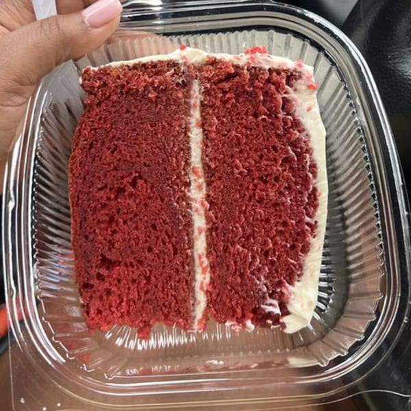 A hand holds a clear plastic container with a slice of red velvet cake with white frosting.